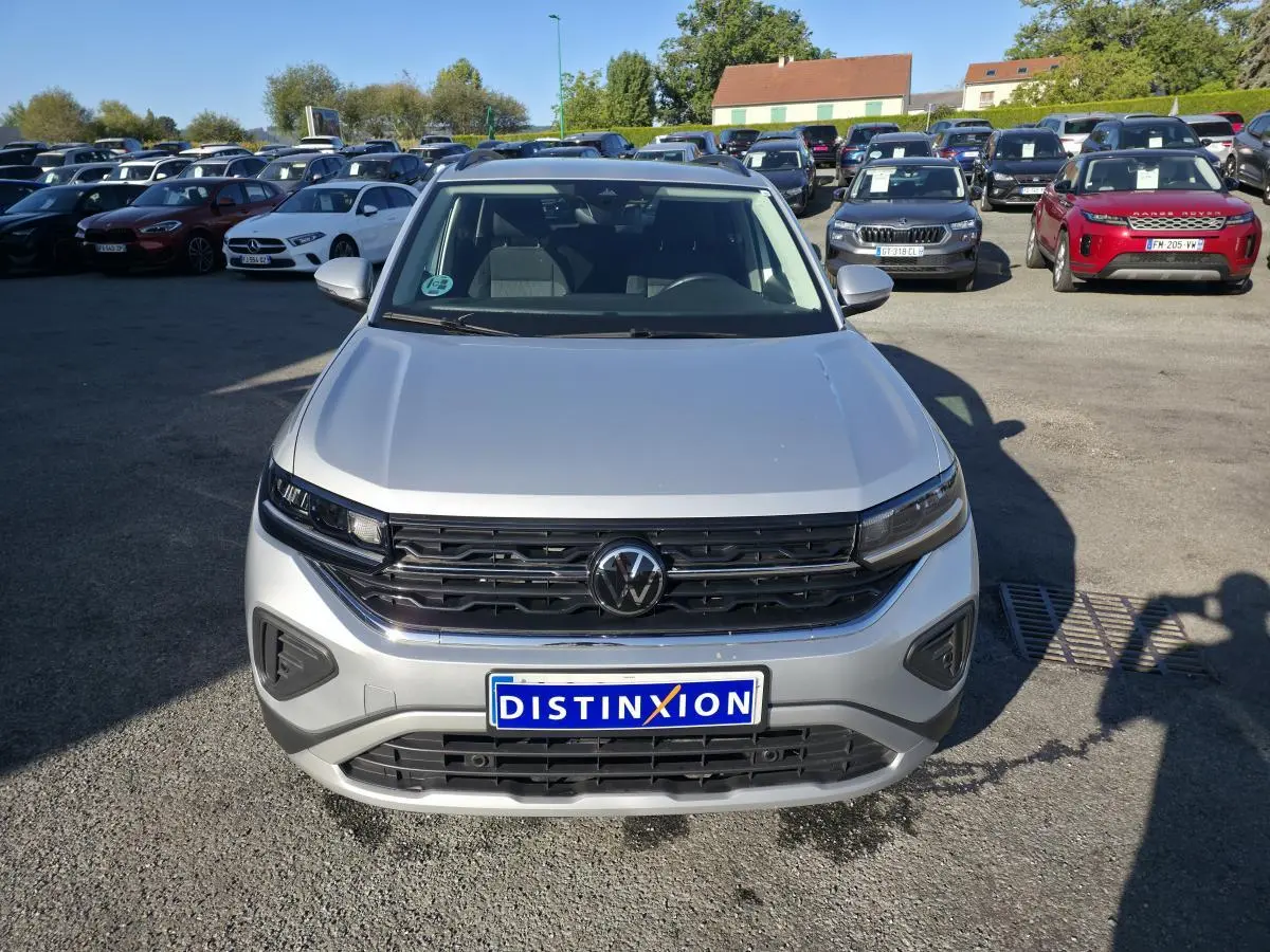 Vue avant d'un Volkswagen T-Cross gris métallisé 2025 avec calandre noire et logo VW central, sur parking extérieur.