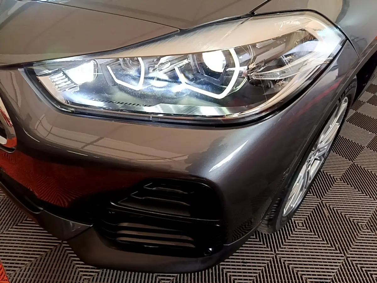 Gros plan sur le phare avant droit allumé du BMW X2 xDrive25e gris métallisé avec jante visible sur sol à motif.