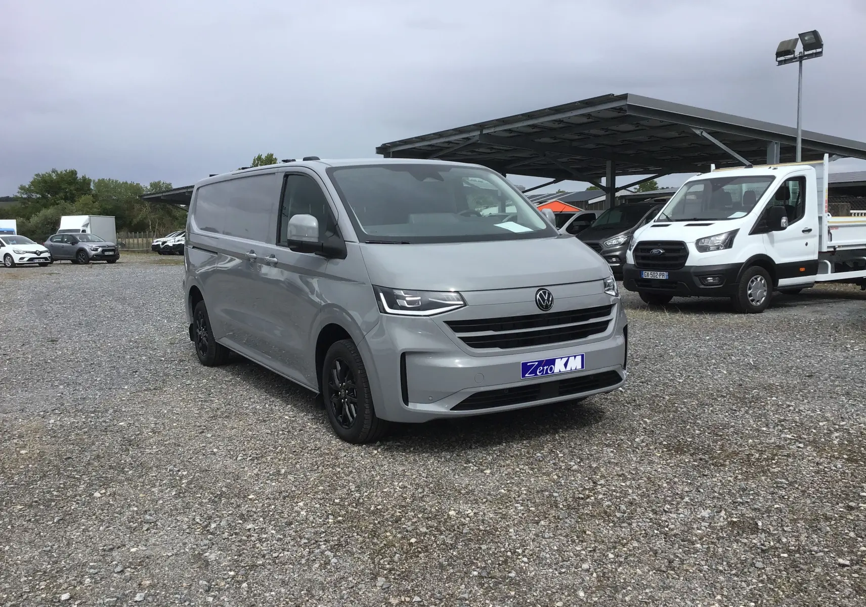 Vue 3/4 avant droit d’un Volkswagen Transporter T7 gris stone avec jantes noires sur un parking extérieur.