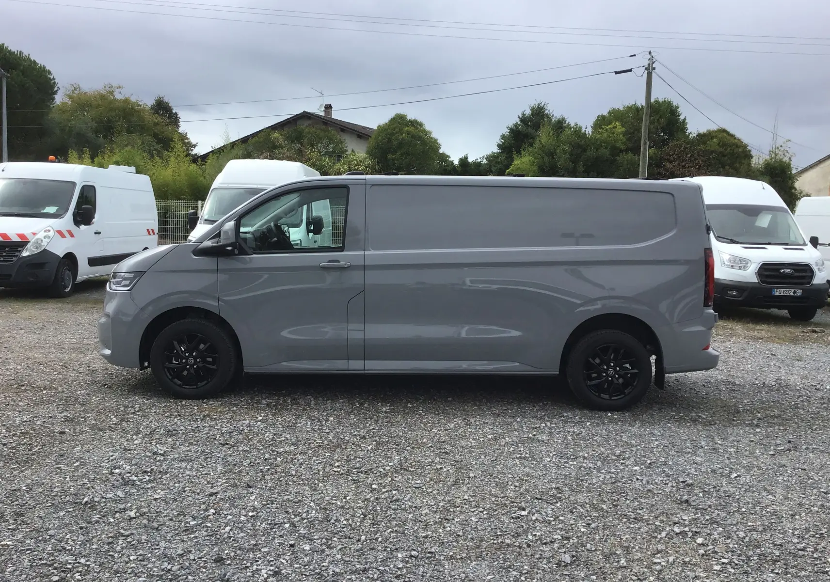 Profil côté gauche d'un Volkswagen Transporter T7 gris stone avec jantes noires sur un parking extérieur.