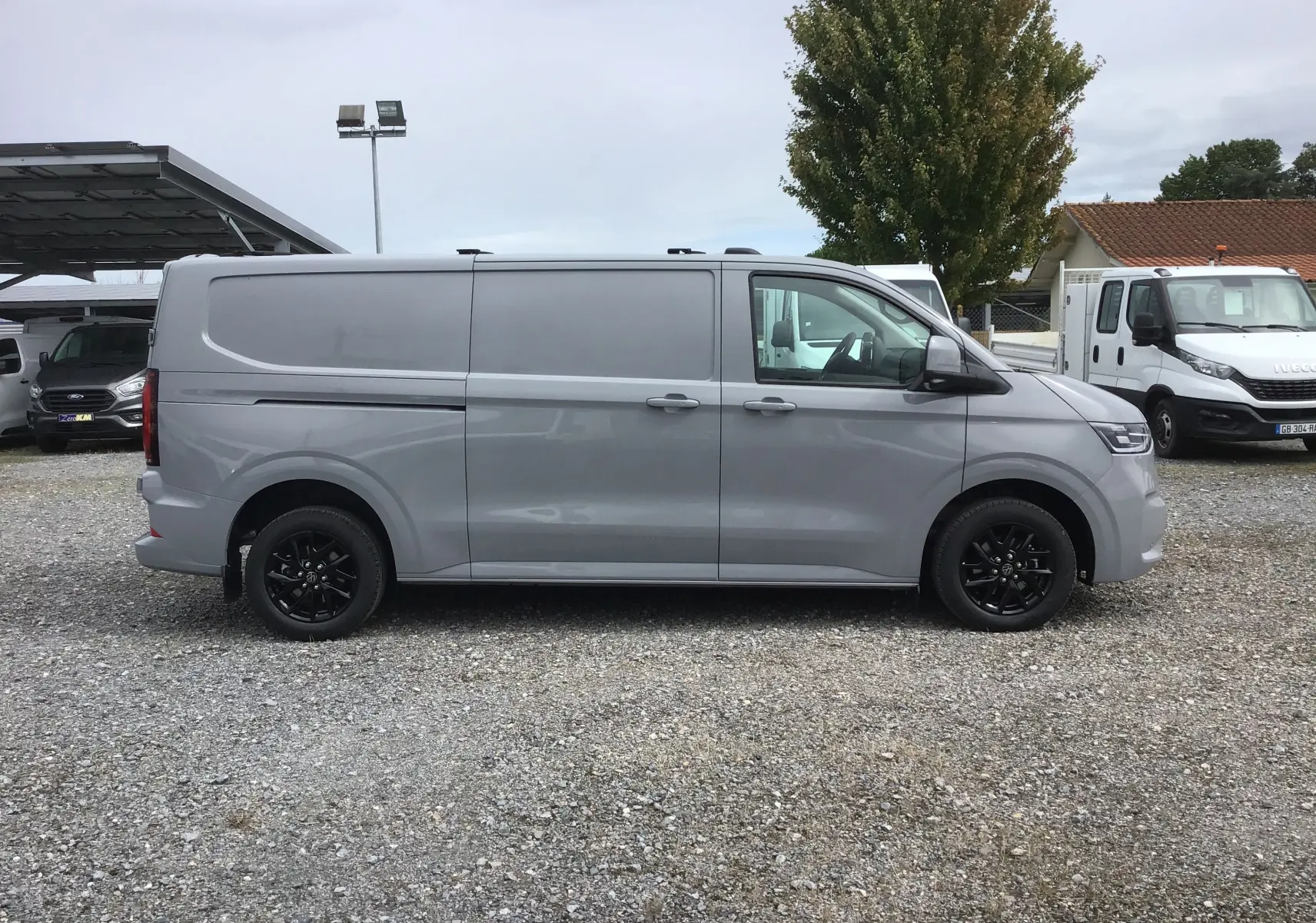 Vue de profil côté gauche d'un Volkswagen Transporter T7 gris stone avec jantes noires et barres de toit repliables.