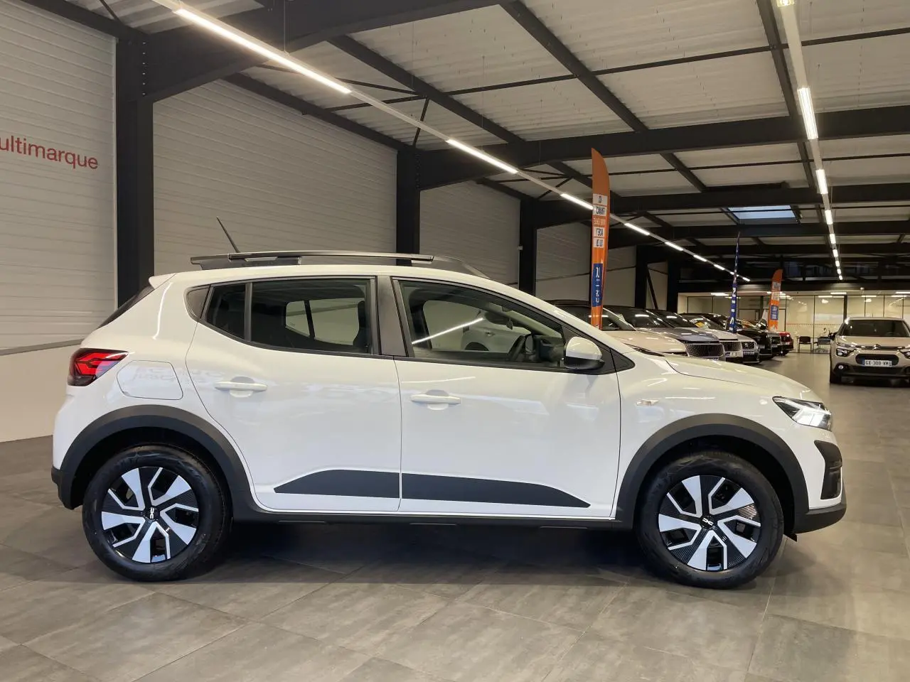 Vue latérale droite d'une Dacia Sandero Stepway blanche Glacier avec barres de toit et jantes 16 pouces en concession.