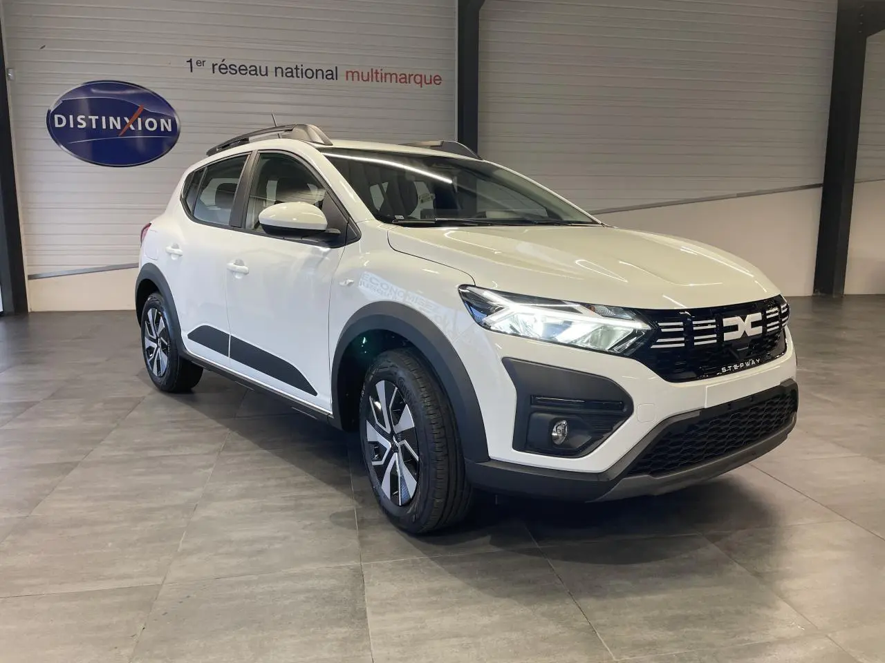 Vue 3/4 avant d'une Dacia Sandero Stepway Expression 2025 blanc glacier avec jantes spécifiques et barres de toit.