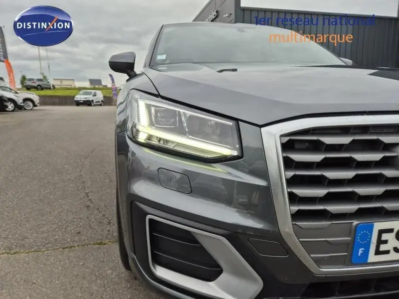 Gros plan sur l'avant gauche d'un Audi Q2 gris Manhattan avec phares LED allumés et calandre hexagonale.