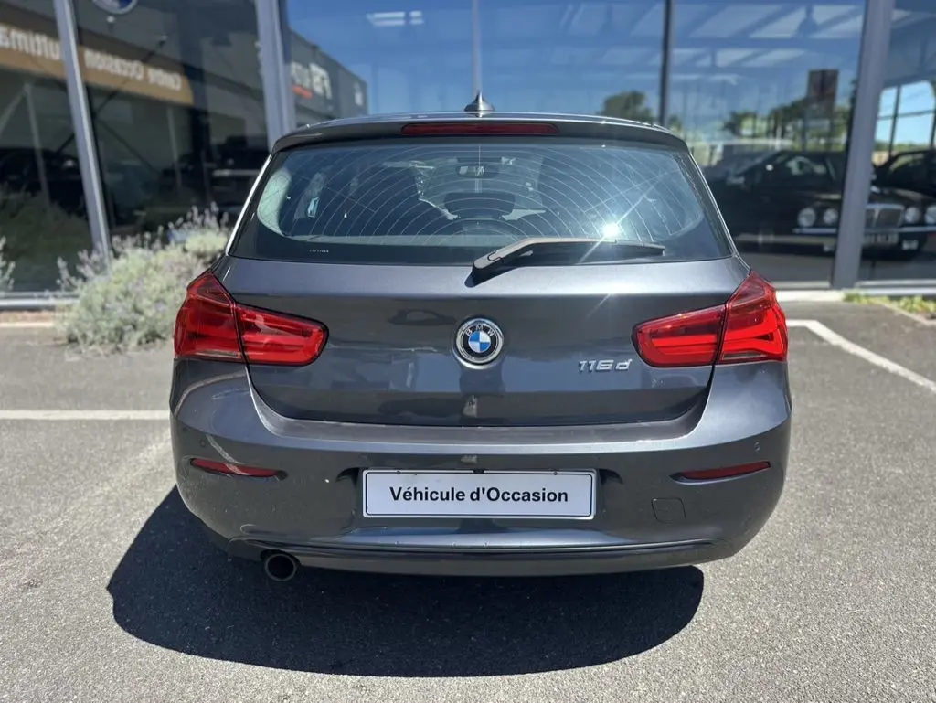 Vue arrière d'une BMW Série 1 F20 gris foncé avec logo et badge 116d, stationnée en extérieur devant un showroom.