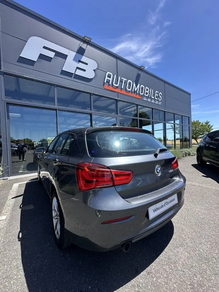 BMW Série 1 F20 gris foncé vue 3/4 arrière droit, avec feu arrière rouge et plaque "Véhicule d'Occasion" visible.