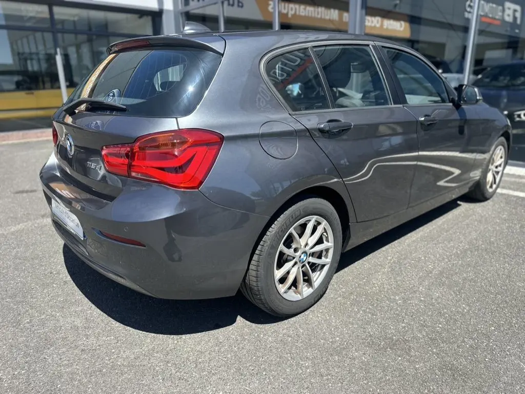 BMW Série 1 F20 gris foncé vue 3/4 arrière droit, mettant en valeur les feux arrière et les jantes alliage.