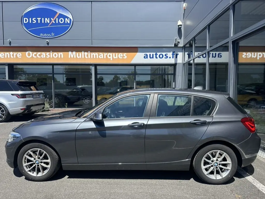 Profil côté gauche d'une BMW Série 1 F20 gris foncé, modèle 116D Business Design 2019, garée devant un showroom.