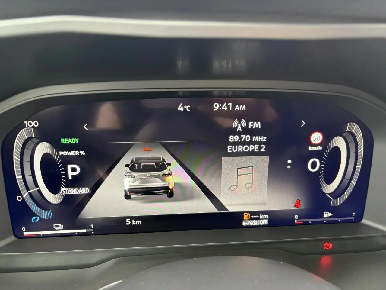 Affichage numérique du tableau de bord du Nissan Qashqai hybride, montrant la vitesse, la radio et l'icône du véhicule en vue arrière.