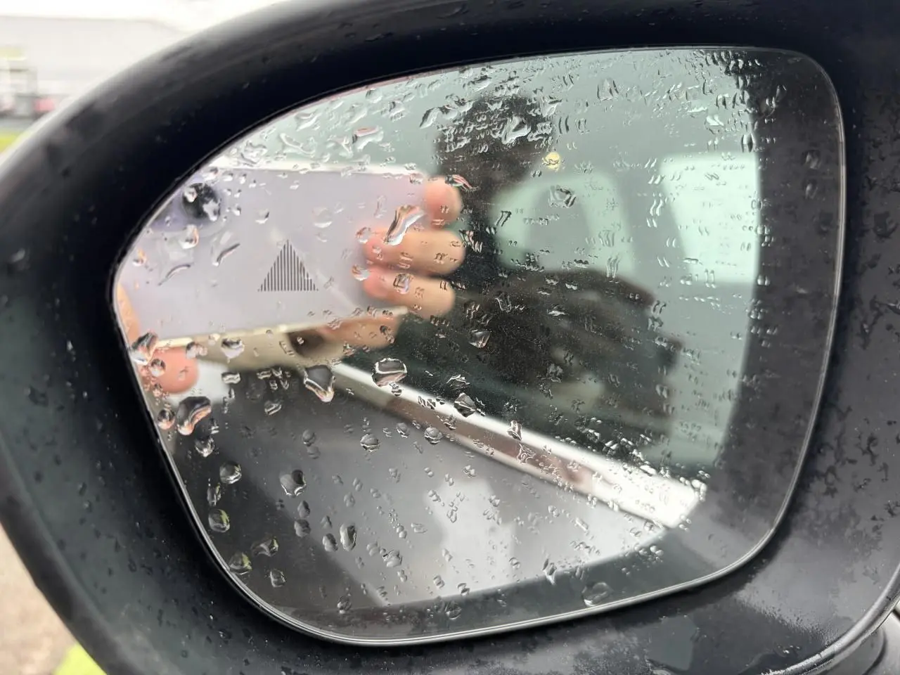 Rétroviseur extérieur droit noir avec gouttes d'eau sur un Nissan Qashqai gris foncé, reflet flou d'une main et d'un véhicule.