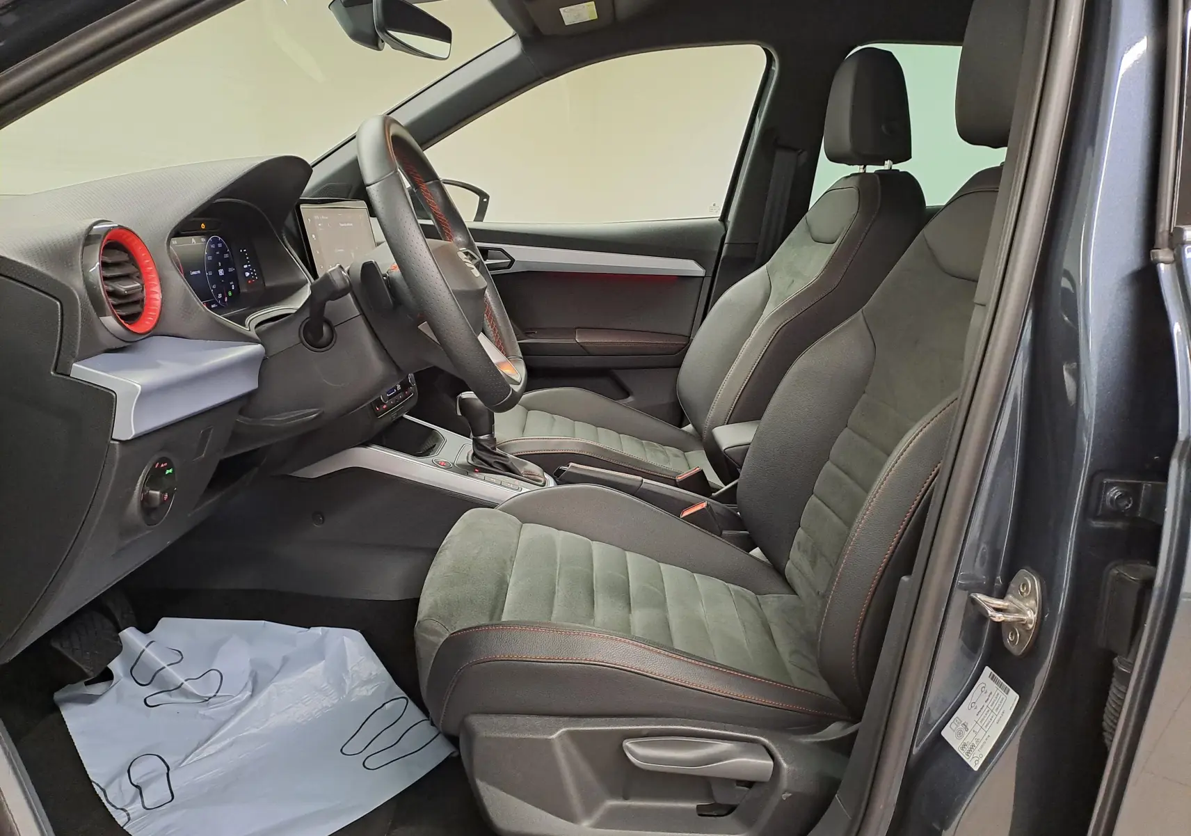 Vue intérieure côté conducteur du SEAT Arona 2024 gris magnétique, sièges sport gris avec surpiqûres orange et tableau de bord moderne.