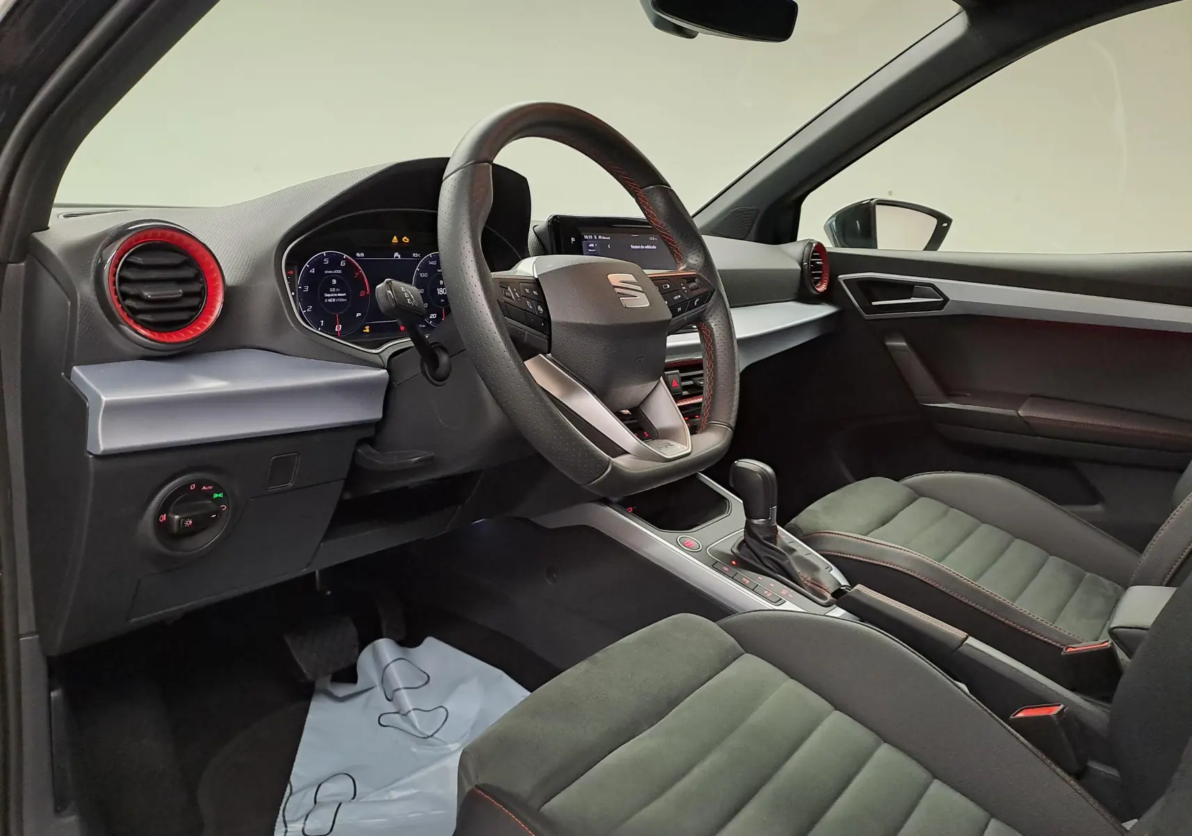 Vue intérieure côté conducteur du SEAT Arona 2024, volant cuir, aérateurs rouges et sièges tissu gris avec surpiqûres rouges.