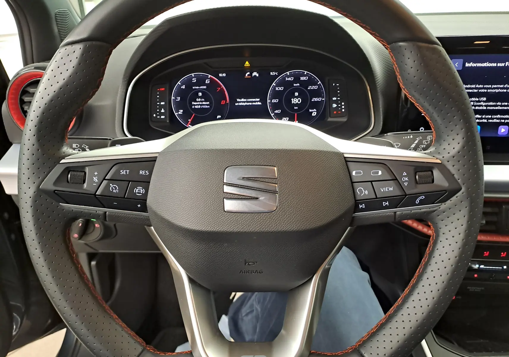 Vue rapprochée du volant perforé avec surpiqûres orange du SEAT Arona 2024, tableau de bord numérique en arrière-plan.