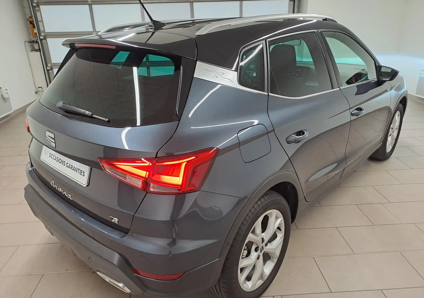 Vue 3/4 arrière droit d'un SEAT Arona 2024 gris magnétique avec toit noir et feux arrière allumés en intérieur.