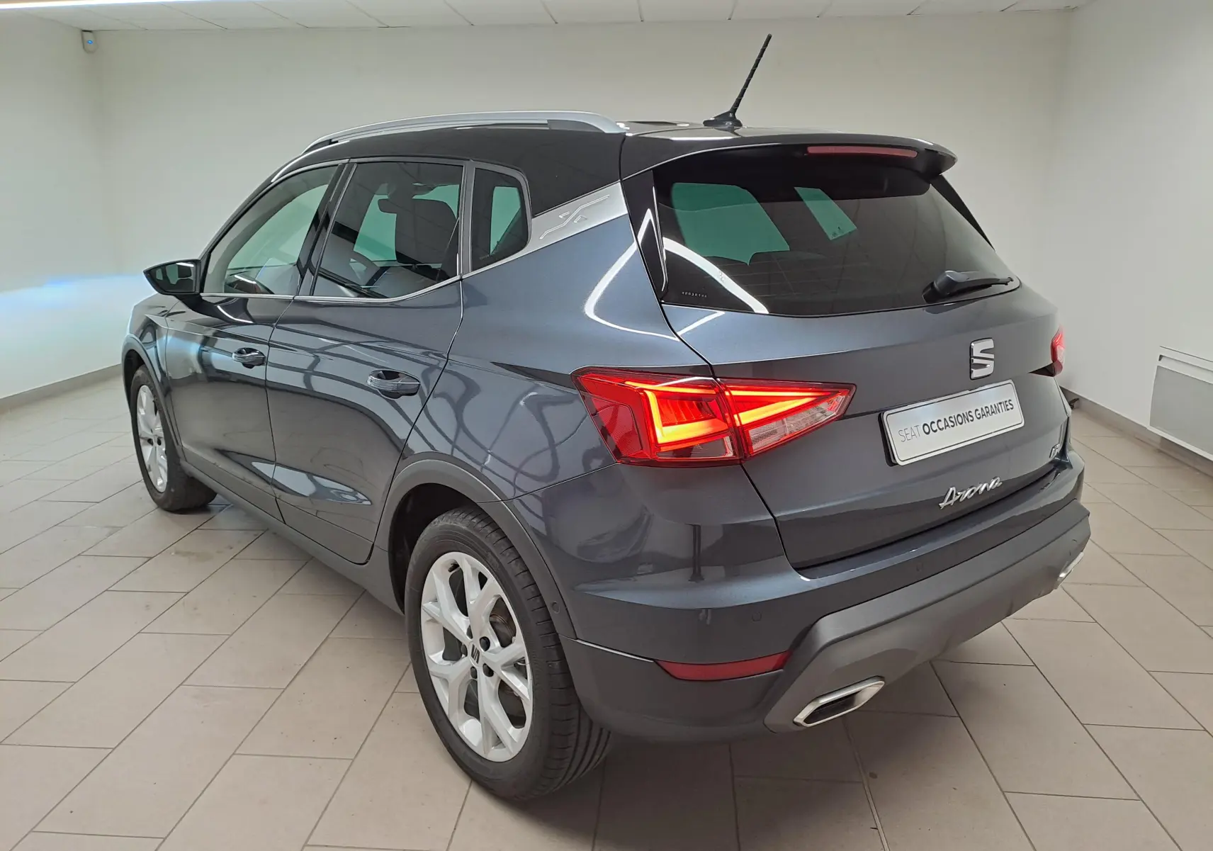 Vue 3/4 arrière droite du SEAT Arona 2024 gris magnétique avec toit noir et feux arrière allumés dans un intérieur neutre.