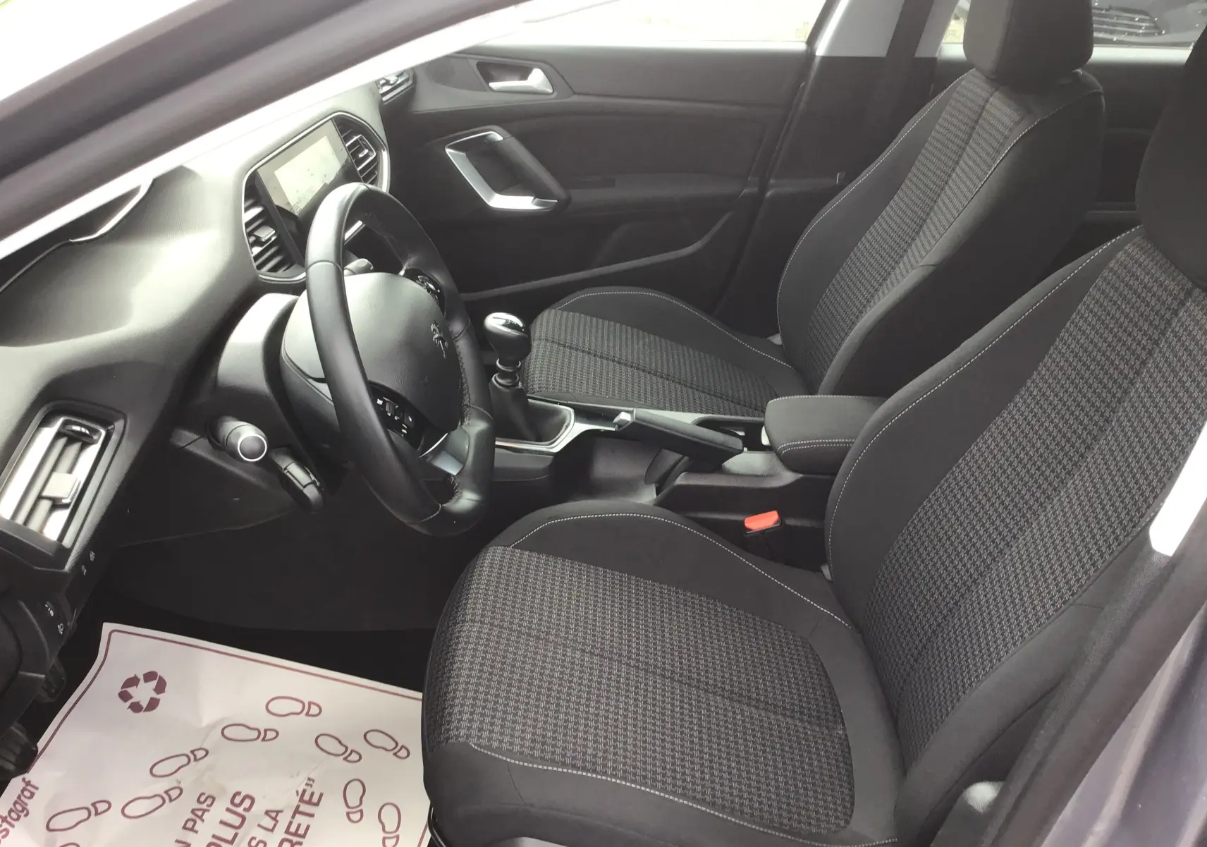 Vue intérieure côté conducteur de la Peugeot 308 1.5 BlueHDi gris Artense, sièges tissu à motif pied-de-poule et boîte manuelle.