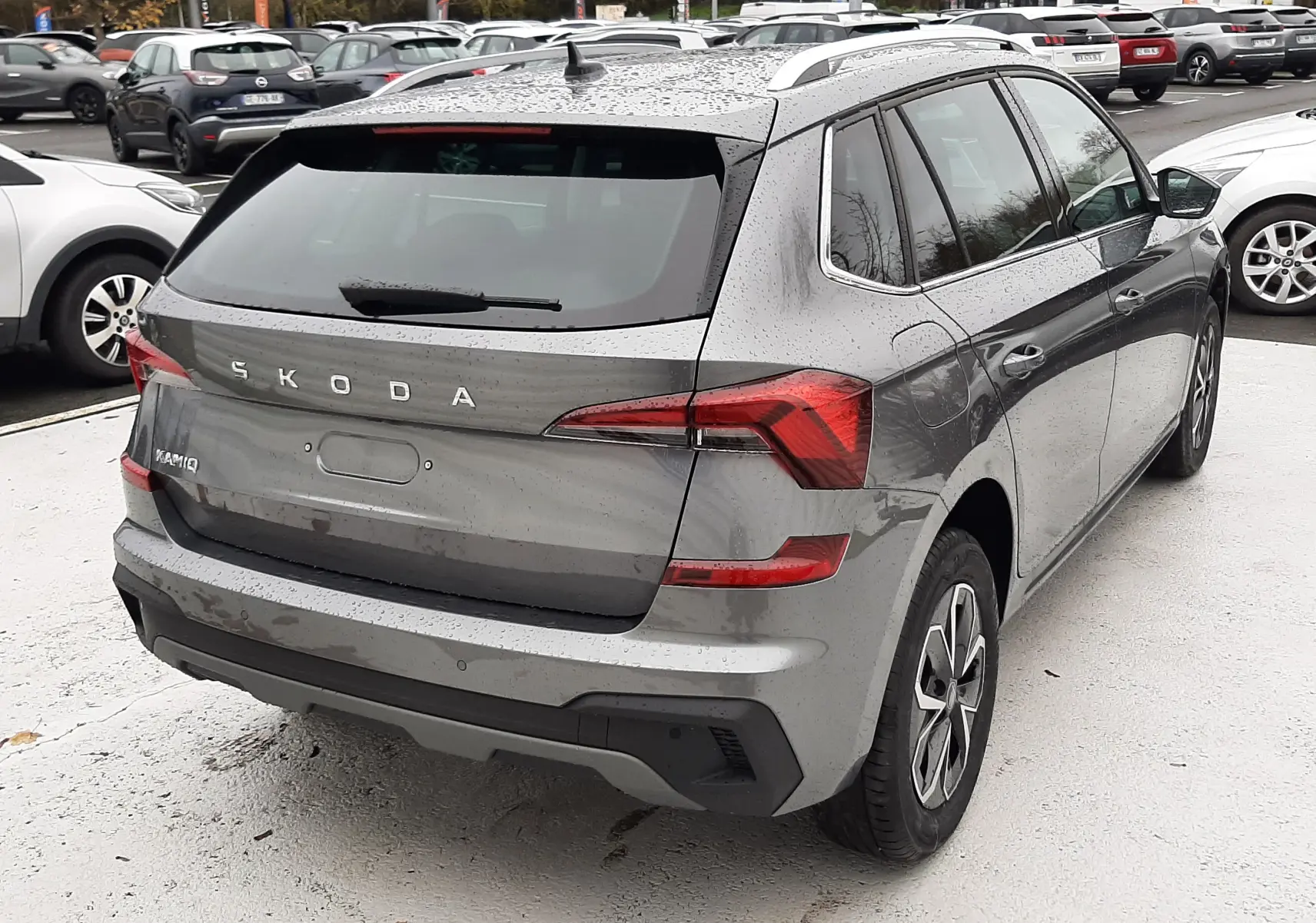 Vue 3/4 arrière droite d'un Skoda Kamiq gris graphite métallisé 2025 avec feux LED et toit avec barres de toit.