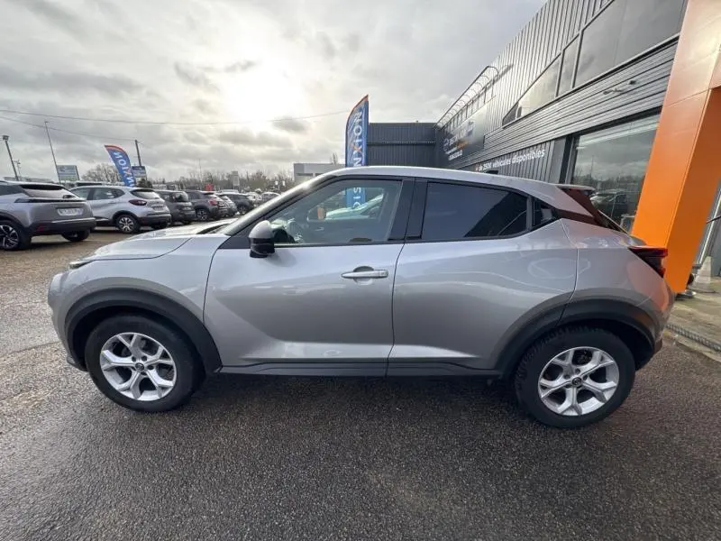 Vue de profil côté gauche d'un Nissan Juke gris perle métallisé garé devant un bâtiment commercial.