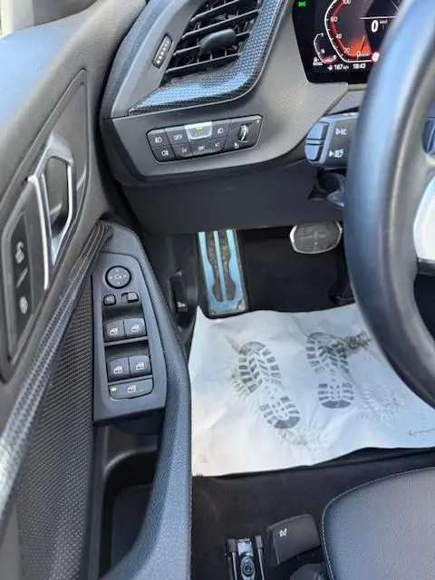 Vue intérieure côté conducteur de la BMW Série 1 118d Edition Sport 2024, avec commandes de vitres et pédalier alu.