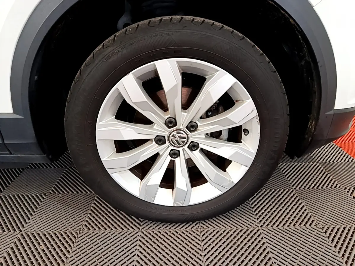 Gros plan sur la roue avant gauche avec jante argentée du Volkswagen T-Roc blanc, détail du pneu et logo VW visible.
