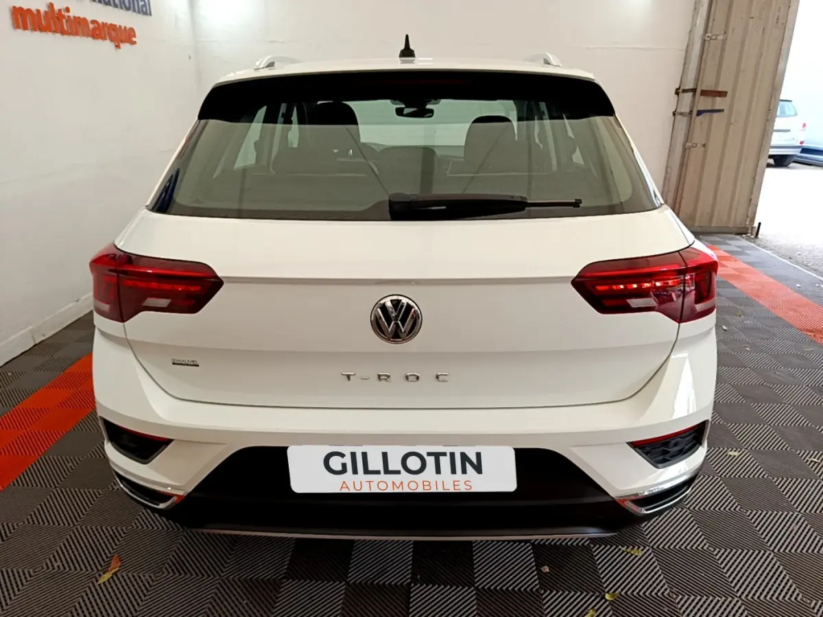 Vue arrière d'un Volkswagen T-Roc blanc avec feux arrière allumés et logo VW bien visible.