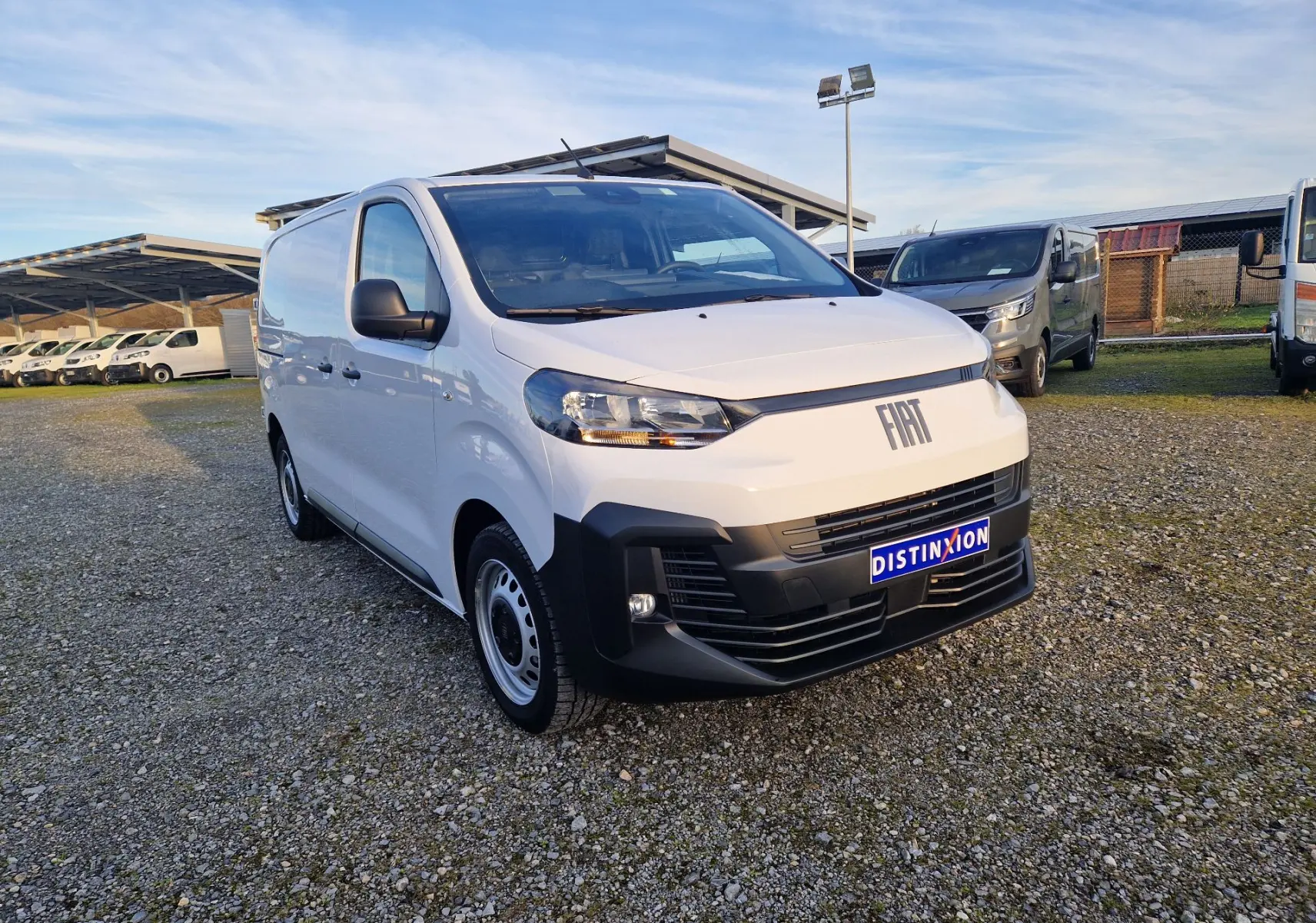 Fiat Scudo Fourgon blanc en 3/4 avant droit, avec pare-chocs noir et plaque de protection, stationné sur un sol gravillonné.
