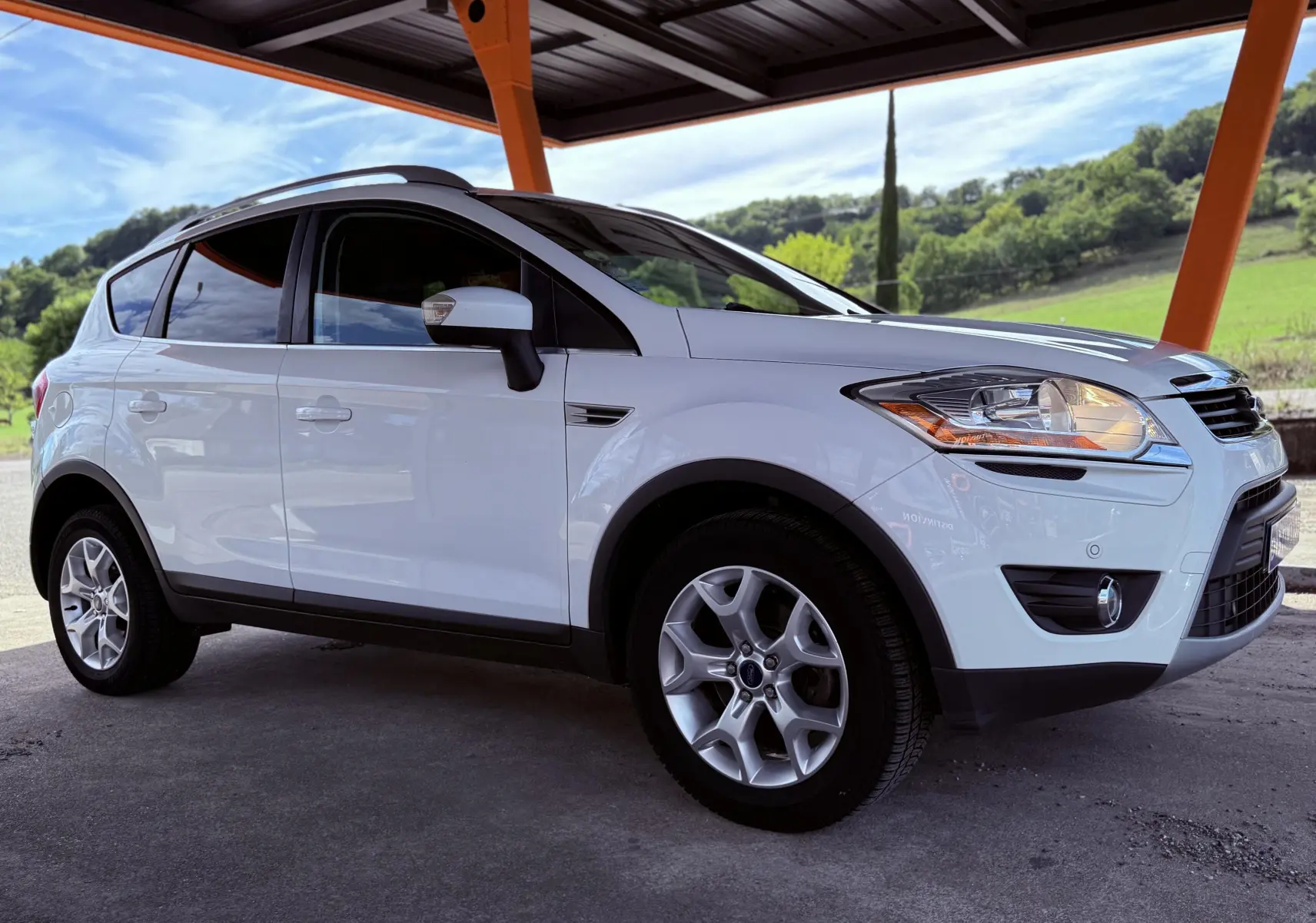 Ford Kuga blanc vu de profil côté gauche sous un abri avec un paysage verdoyant en arrière-plan.