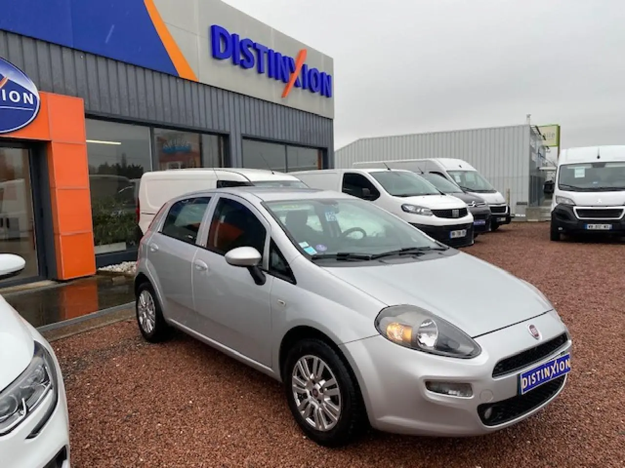 Fiat Grande Punto gris métallisé vue 3/4 avant droit avec phares allumés sur parking de concession.