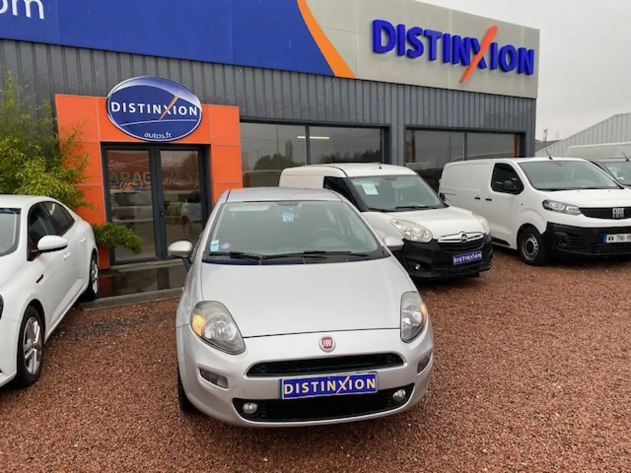 Vue avant d'une FIAT Grande Punto gris métallisé stationnée devant un showroom Distinxion par temps couvert.