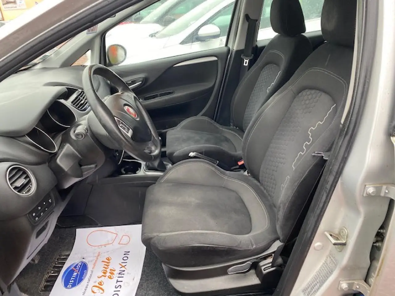 Vue intérieure côté conducteur de la Fiat Grande Punto 1.2i gris, montrant les sièges noirs et le volant multifonction.