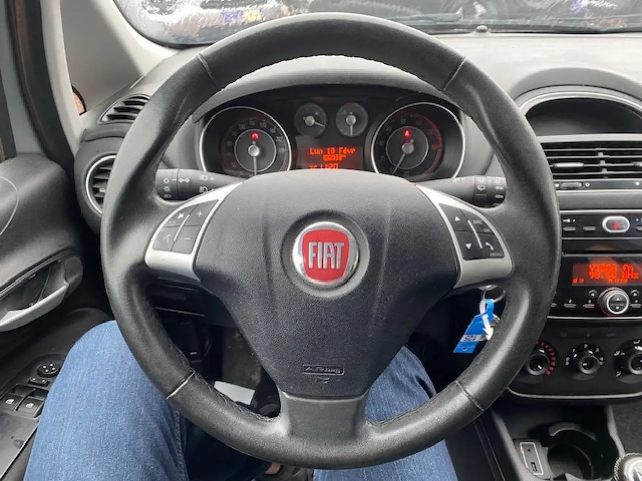 Vue intérieure centrée sur le volant noir de la Fiat Grande Punto 2015 avec commandes audio intégrées et tableau de bord visible.