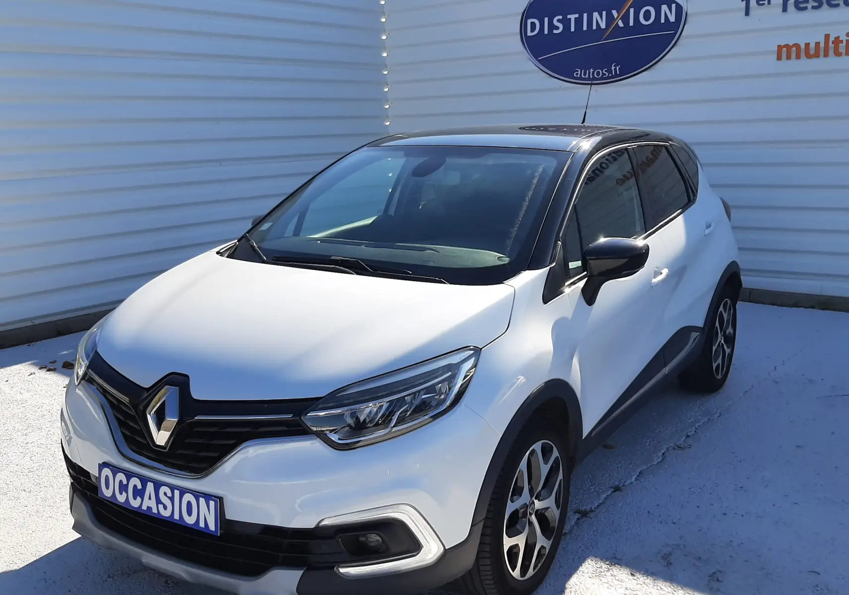 Renault Captur blanc nacré avec toit noir, vue 3/4 avant droit, sur parking devant un mur blanc.