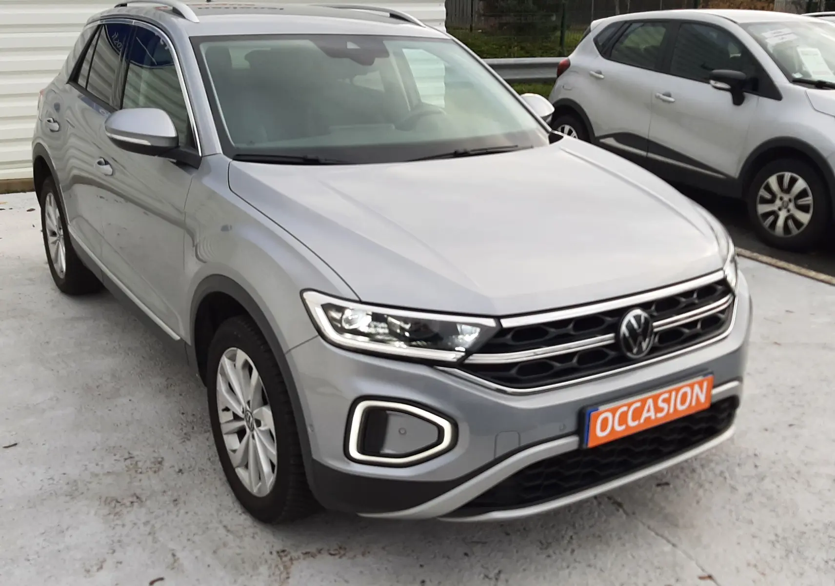 Vue frontale d'un Volkswagen T-Roc gris pyrite 2023 avec calandre noire et feux LED allumés, sur fond de mur blanc.