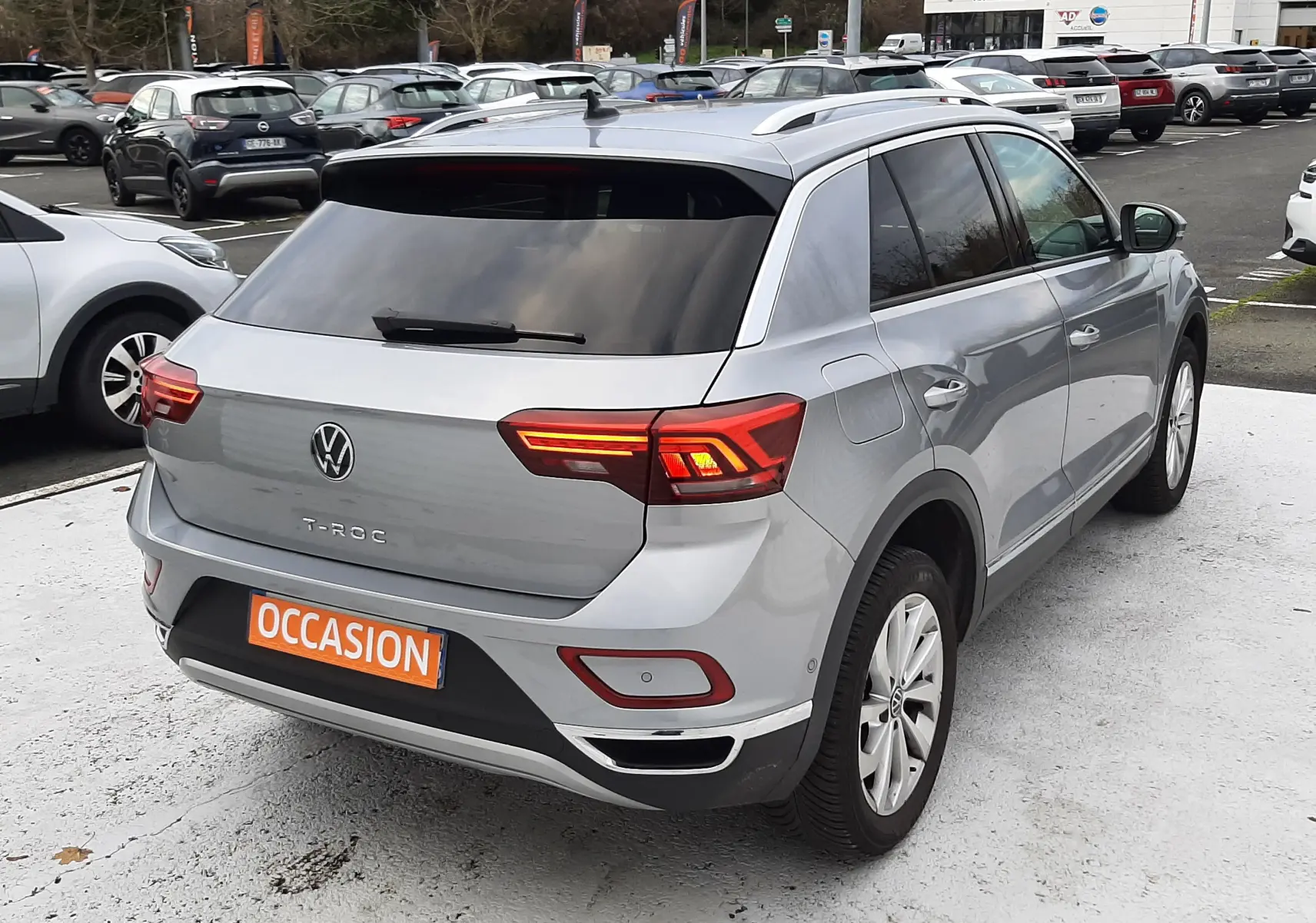 Vue arrière d'un Volkswagen T-Roc gris pyrite 2023 avec feux LED allumés et plaque "OCCASION" orange.
