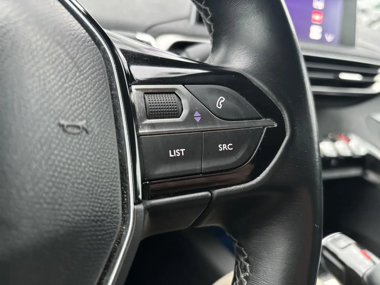 Gros plan sur les commandes gauche du volant noir du Peugeot 3008 blanc, avec écran tactile en arrière-plan.