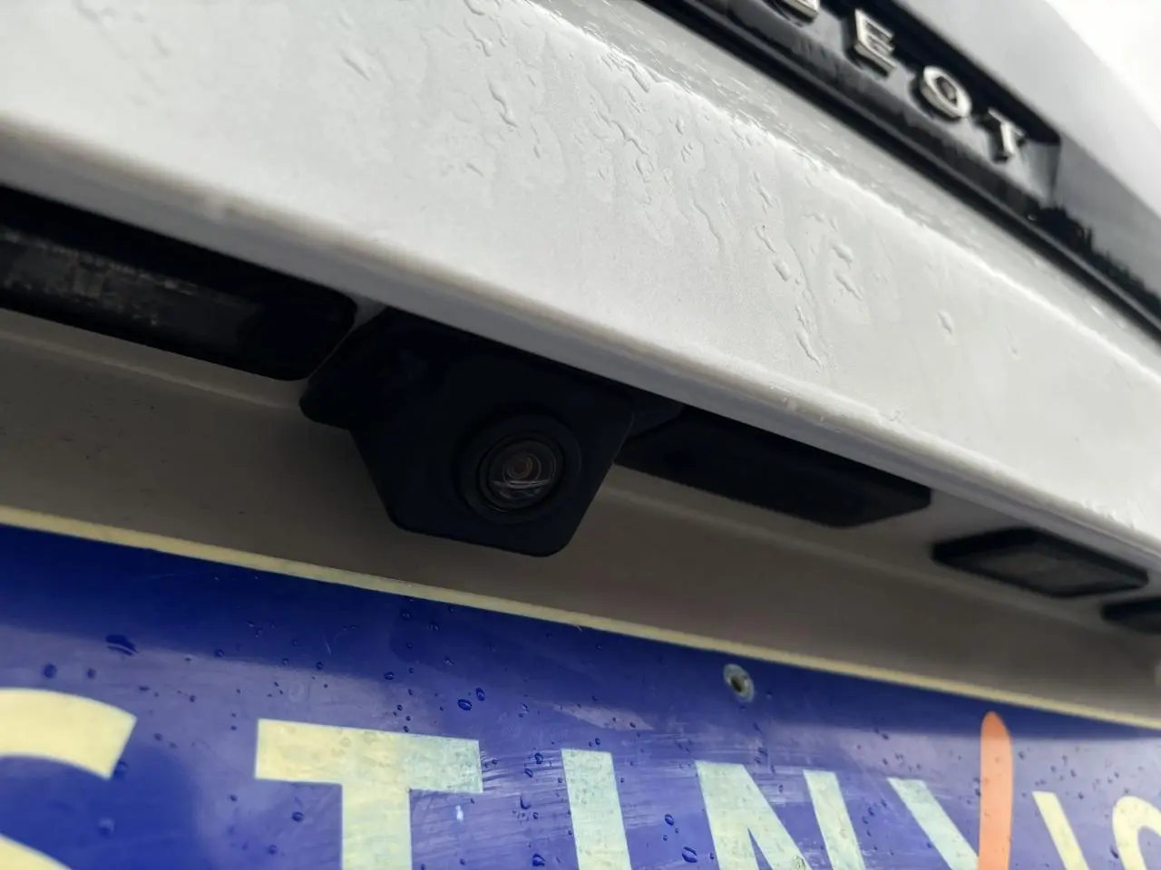 Gros plan sur la caméra de recul noire intégrée au hayon blanc nacré du Peugeot 3008, au-dessus de la plaque d'immatriculation.