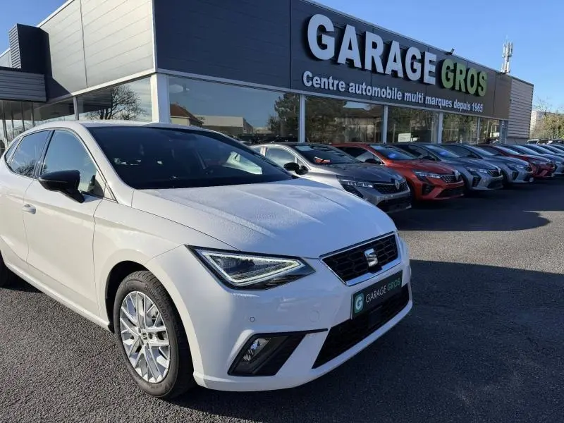 Vue 3/4 avant gauche d'une SEAT Ibiza 1.0 TSI 115cv FR blanche Nevada métal stationnée devant un garage multimarques.