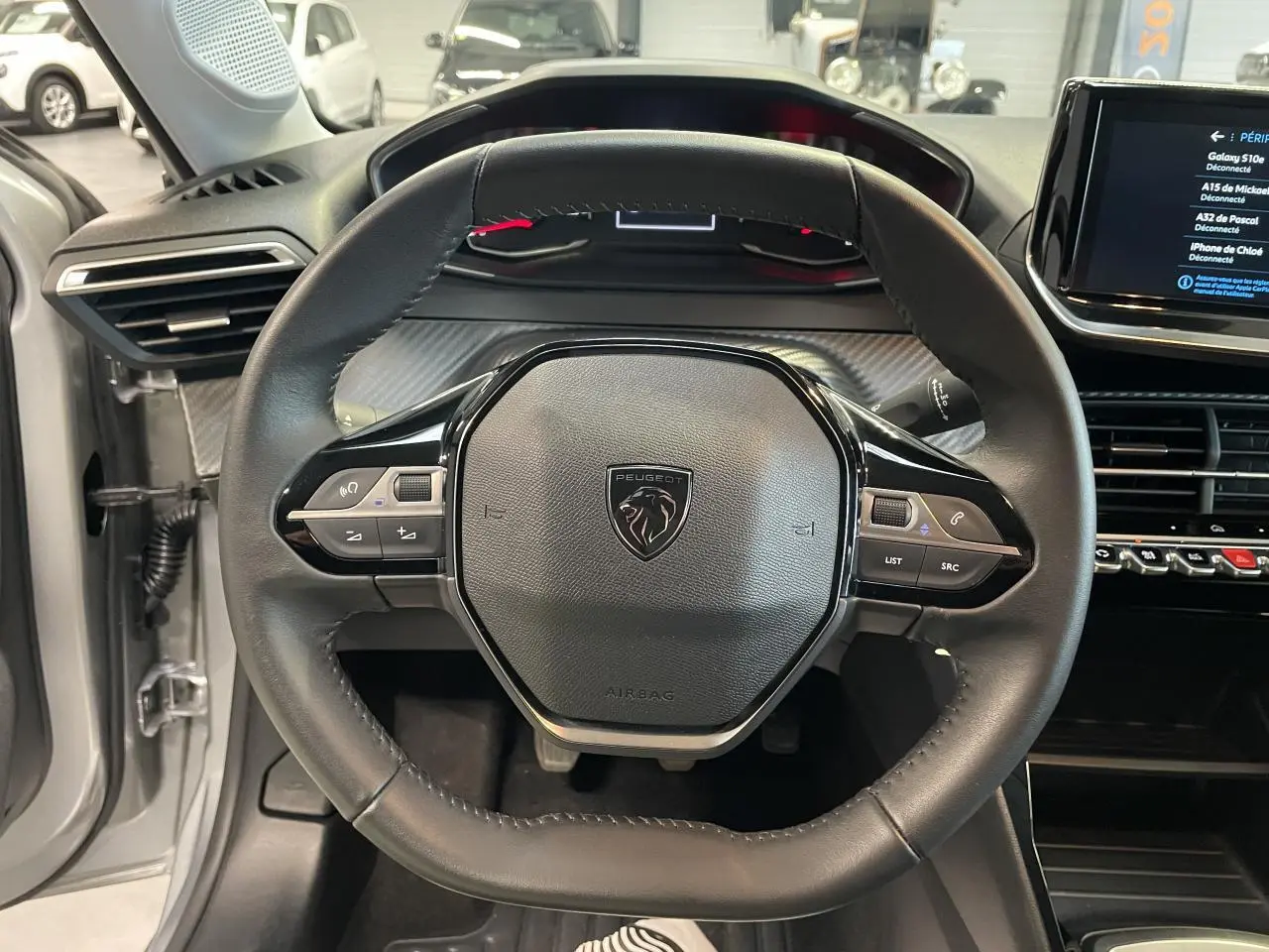 Vue rapprochée du volant cuir noir avec commandes intégrées du Peugeot 208 gris clair, tableau de bord et écran tactile visibles.