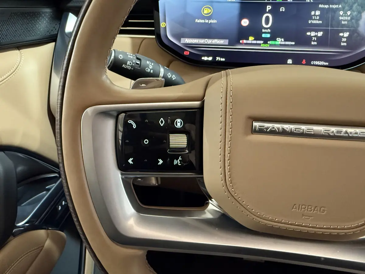 Gros plan sur le volant beige du Range Rover PHEV 2024 avec commandes tactiles et combiné d'instruments numérique visible.