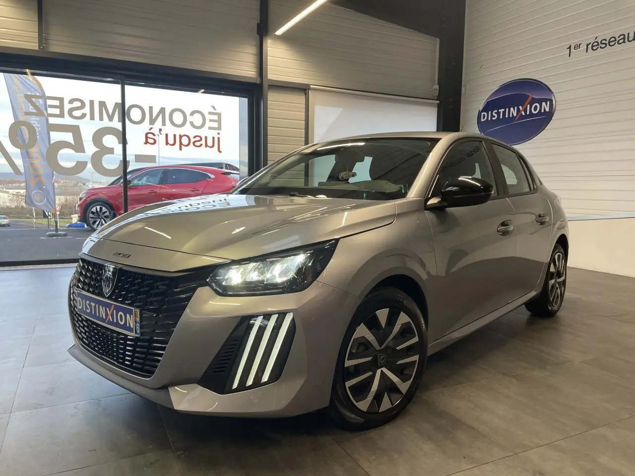Peugeot 208 gris clair en 3/4 avant droit avec feux LED et jantes tôle 16 pouces en showroom.