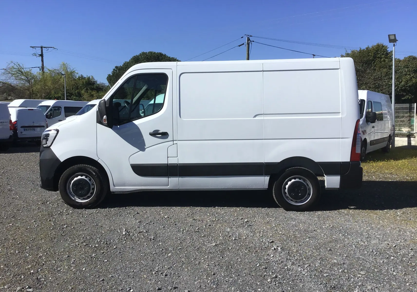 Vue latérale gauche d’un utilitaire Renault Master blanc L1H1 avec bandeau noir et jantes acier, stationné sur sol gravillonné.