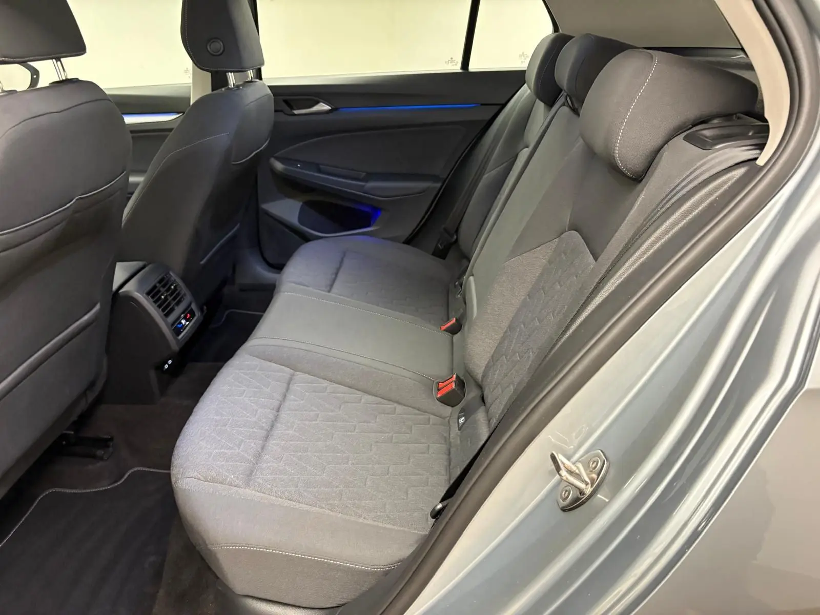 Vue intérieure arrière droite de la Volkswagen Golf 2.0 TDI 2025 en reflet d'argent, sièges tissu gris et éclairage d'ambiance bleu.