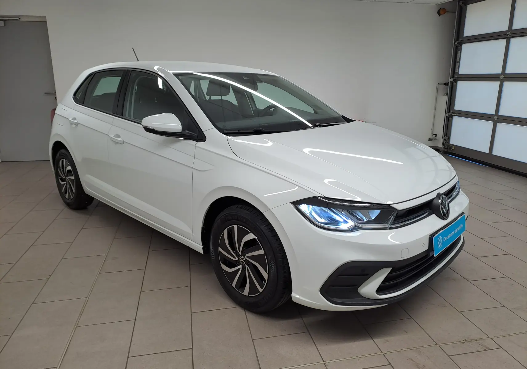 Volkswagen Polo blanc pur vue 3/4 avant droit avec phares allumés dans un garage intérieur.