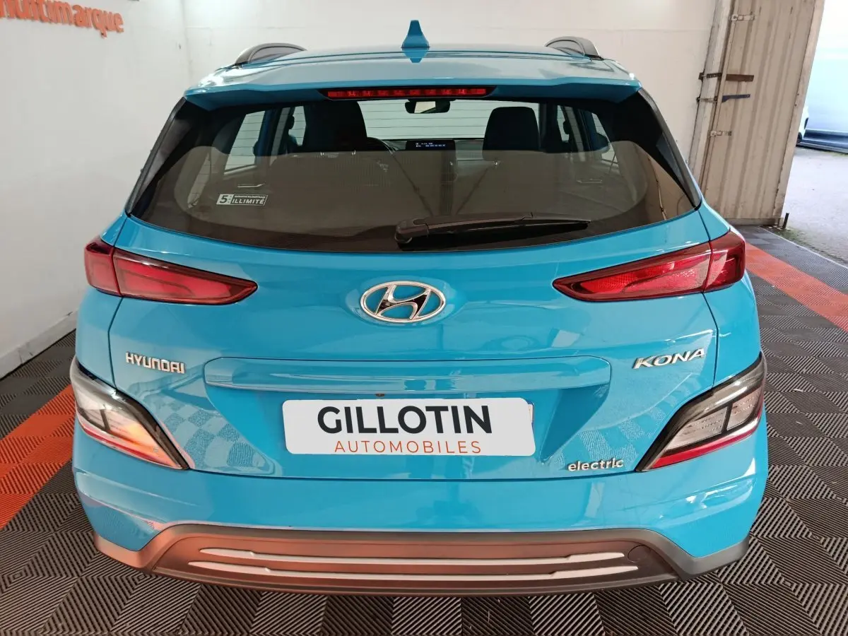 Vue arrière d'une Hyundai Kona électrique bleu vif avec feux arrière allongés et logo visible sur un sol à damier noir et orange.