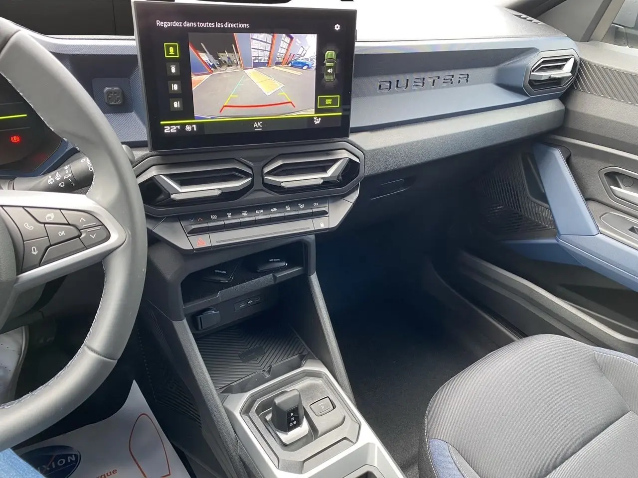 Intérieur du Dacia Duster hybride 2025, tableau de bord noir et bleu avec écran tactile et levier de boîte automatique.