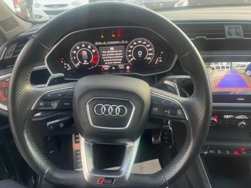 Vue intérieure centrée sur le volant sport perforé noir de l'Audi RS Q3 Sportback avec tableau de bord numérique et écran tactile affichant la caméra de recul.