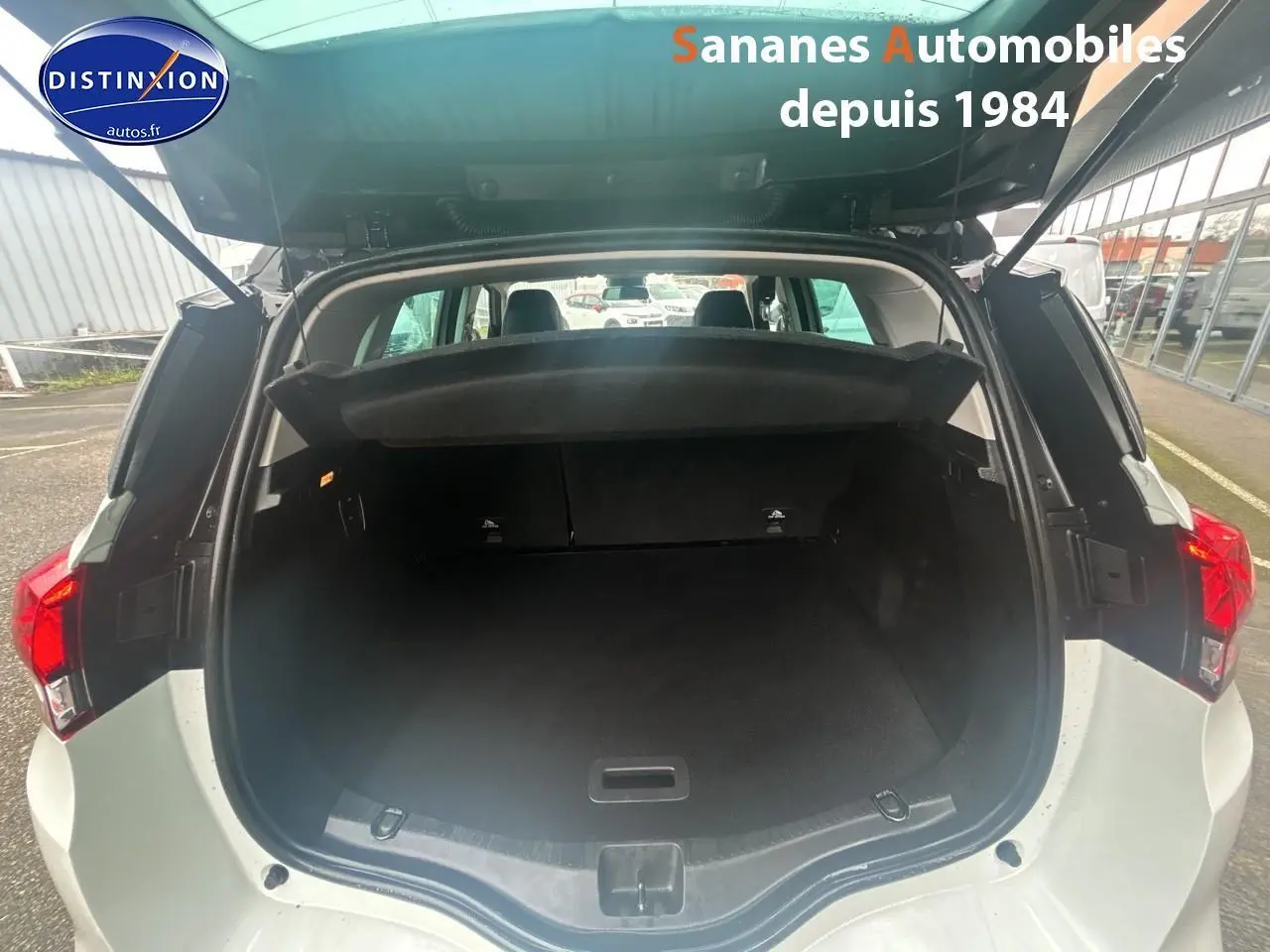 Coffre ouvert vu de l'arrière d'un Renault Scénic blanc nacré avec toit noir étoilé, dans un parking extérieur.