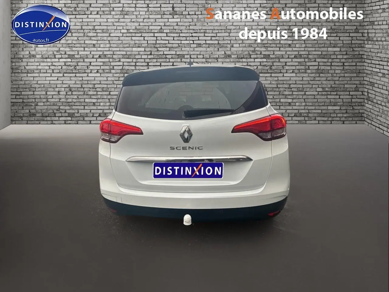 Vue arrière d’un Renault Scénic blanc avec toit noir et attelage, stationné devant un mur en briques grises.