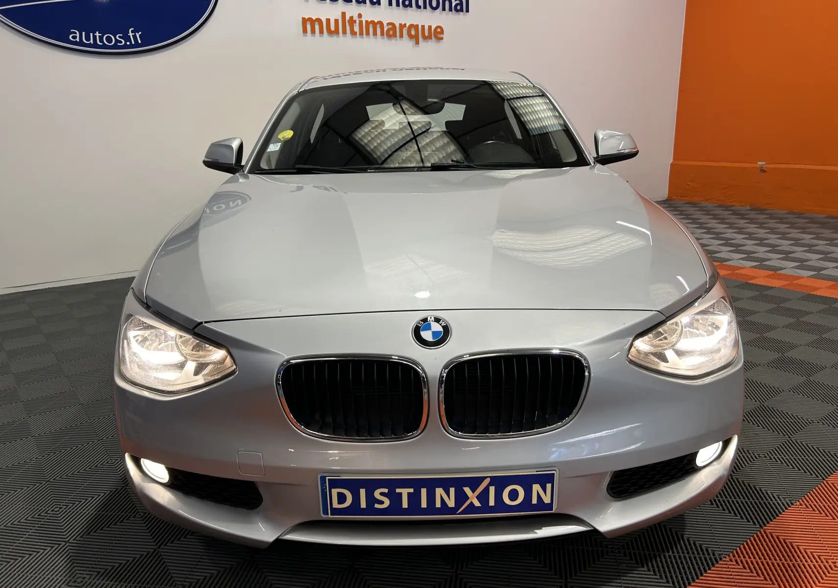 Vue frontale d'une BMW Série 1 gris clair avec phares allumés et plaque Distinxion en concession multimarque.