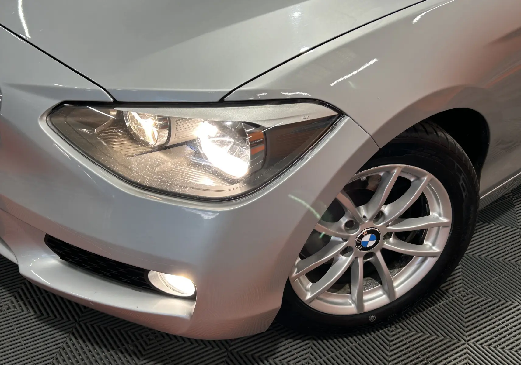Gros plan sur l'avant droit gris d'une BMW Série 1 16d avec phare et jante alliage visible sur sol texturé.